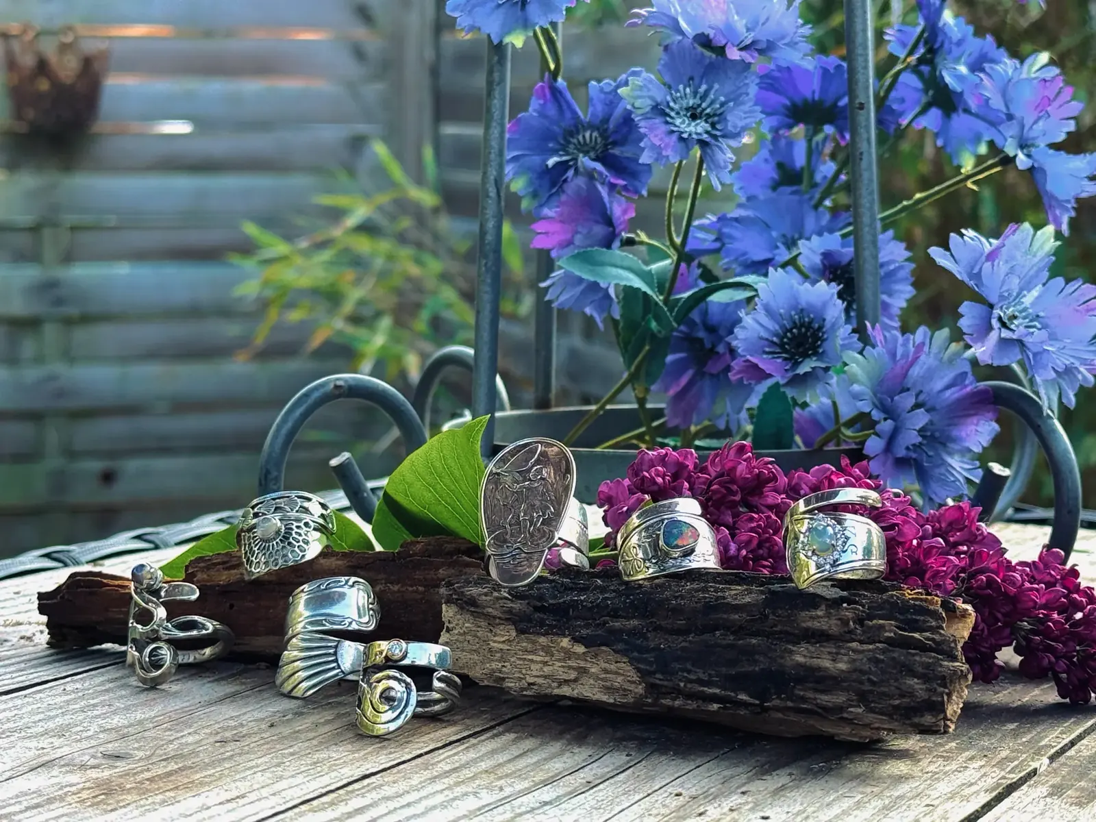 Handgefertigte Ringe aus altem Silberbesteck auf einem Stück Rinde vor Kornblumen